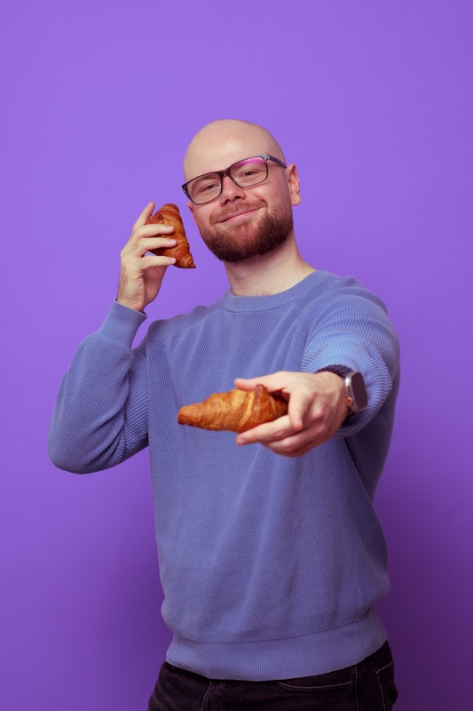 Rubin Koot holds a croissant to his ear like a phone and holds out a second croissant to you