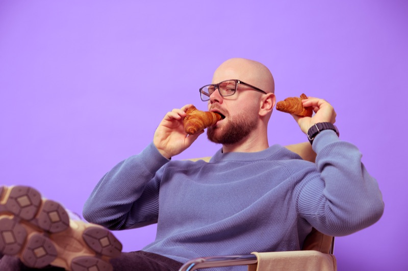 Rubin Koot hangt achterover in een stoel, voeten omhoog, en neemt een hap van een croissant