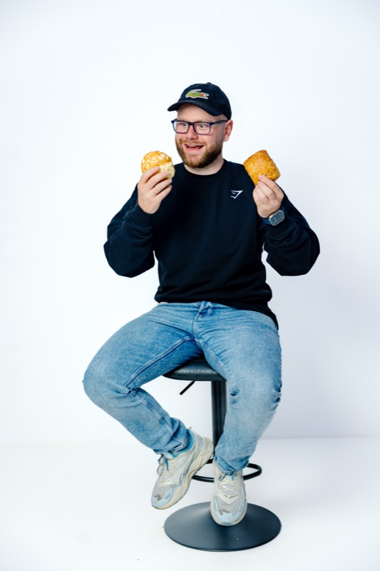 Rubin Koot zit op een barkruk met in elke hand een broodje en lacht